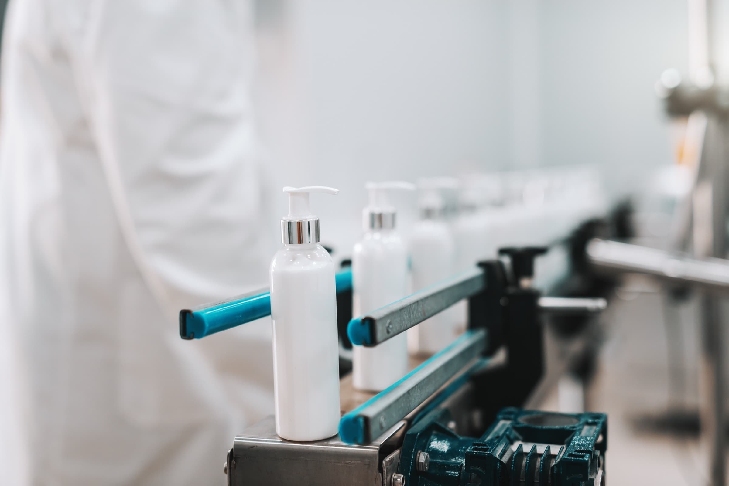 White cosmetic bottles with dosing pumps on a filling line, an operator in a white coat behind them